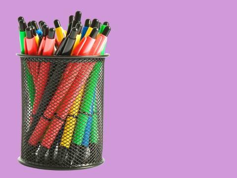 Colorful pens in a metal cup holder, in front of a purple background.