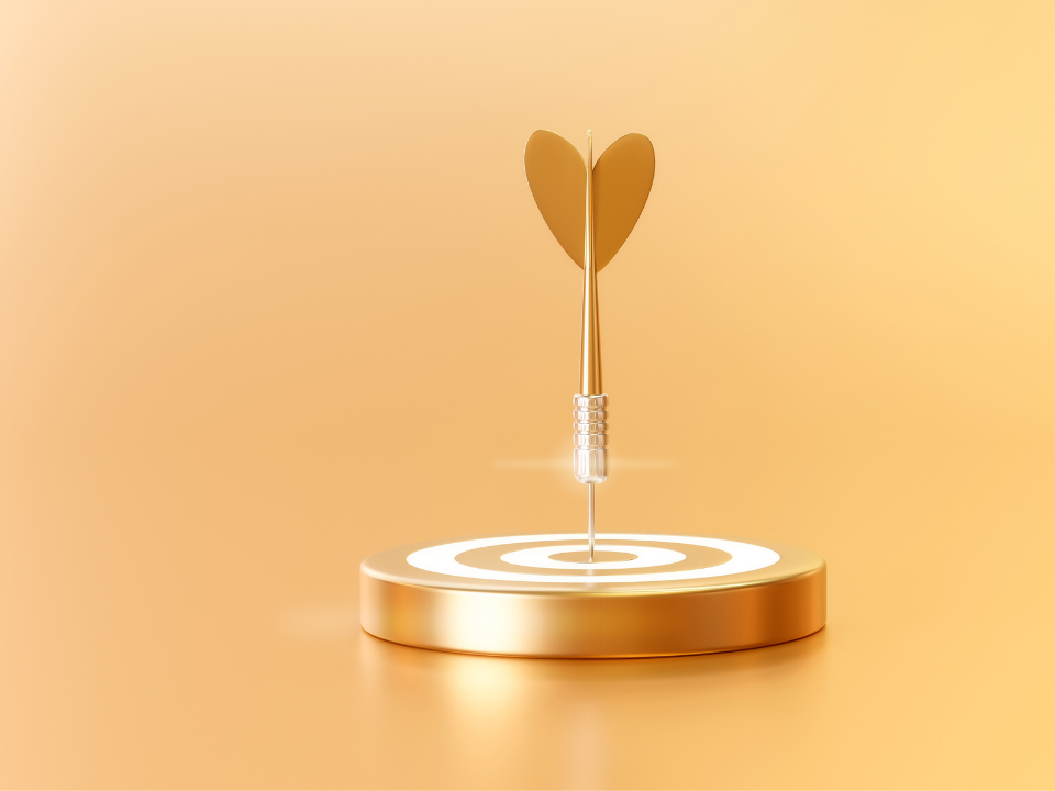 A golden dartboard with a heart shaped dart hitting the bullseye in front of yellow background.
