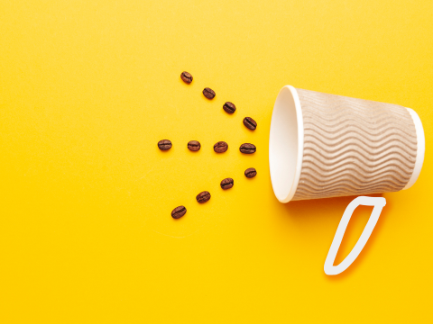 Cup of coffee tipped over with coffee beans coming out of cup in front of a yellow background