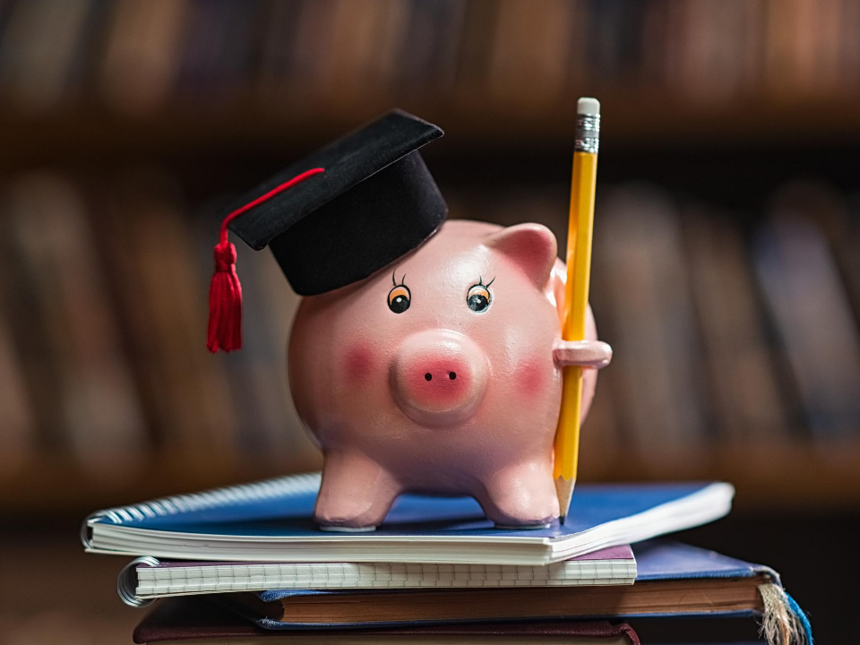 Pink piggy bank with a graduation cap on holding a pencil, sitting on a stack of notebooks